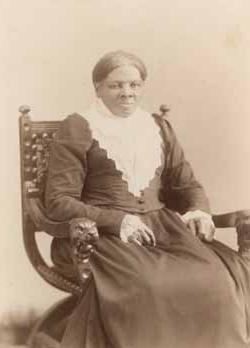 <p>Black and white photograph of a Black woman with her hair parted in the middle and pinned behind her head wearing a black, 长袖连衣裙和三角形蕾丝领. 她坐在一把木椅上，双手放在膝盖上.</p>